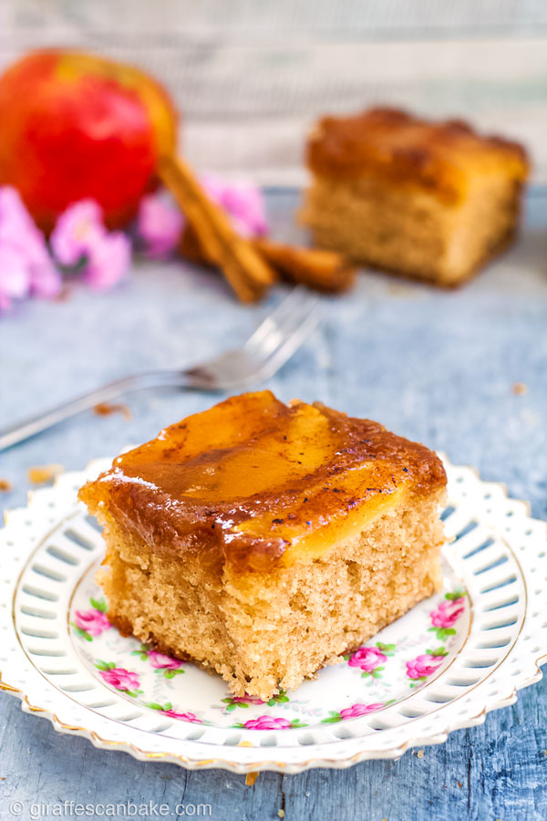 A piece of Gluten Free Apple Upside Down Cake on a white and pink plate, with a whole apple, a forkl, and another piece of cake in the background