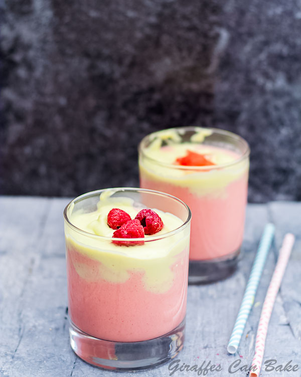 Short glass of citrus smoothie with pink and green layer, topped with raspberries. Another short glass in background - full size